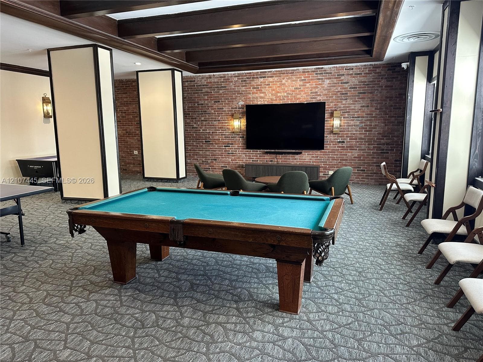 Recreational room featuring exposed brick wall, dark wood-beam ceiling, decorative pillars, patterned carpeting, and built-in wall sconces