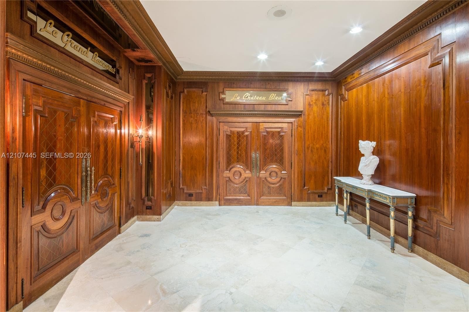 Foyer featuring floor-to-ceiling wood paneling with decorative trim, polished stone flooring, double wood doors with detailed millwork, and wall-mounted sconces