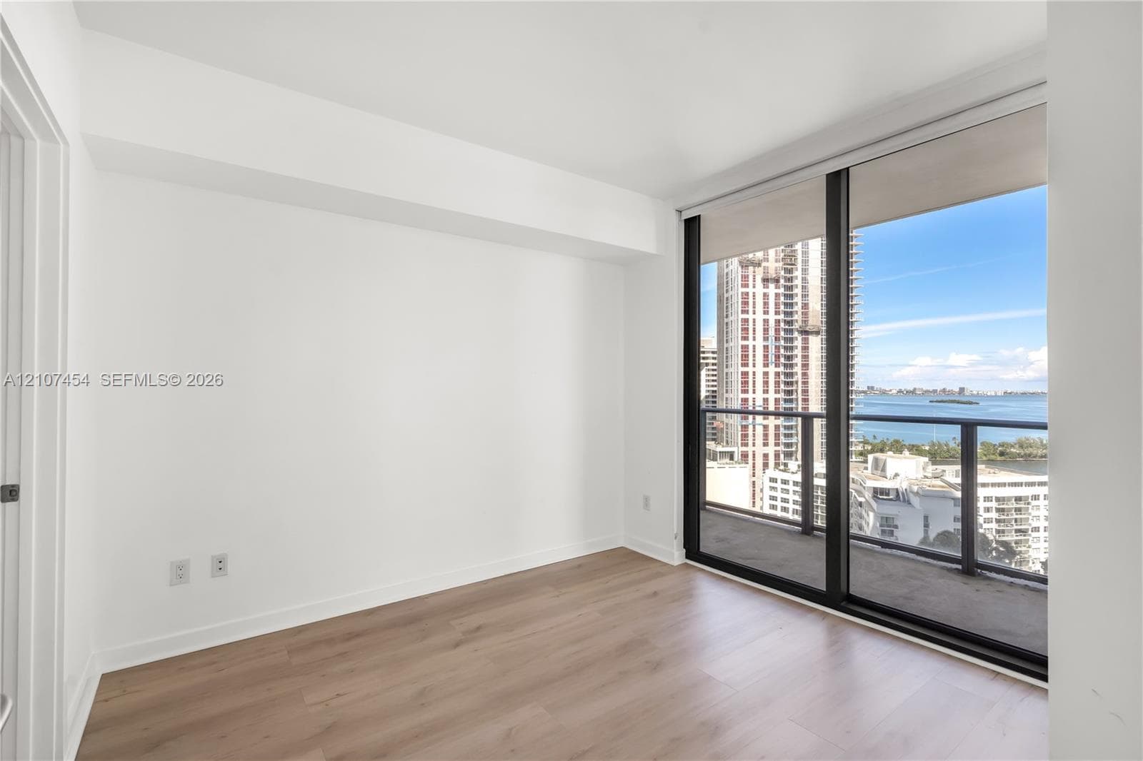 Interior room featuring wood-finish flooring, floor-to-ceiling windows, and access to a private balcony with water and city skyline surroundings