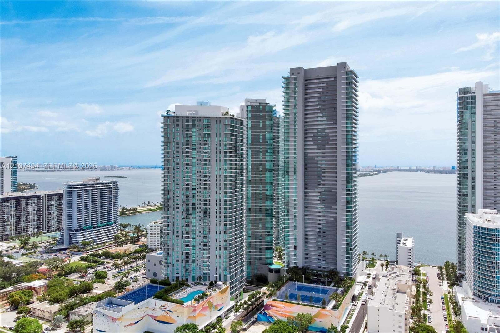 Aerial perspective of a high-rise residential complex featuring glass-facade towers, outdoor swimming pools, and tennis courts situated along the waterfront