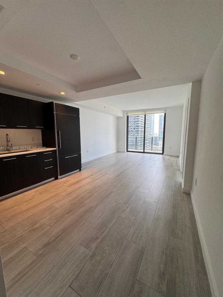 Open-concept living space featuring wood-finish flooring, dark cabinetry, recessed ceiling lighting, and sliding glass doors with balcony access