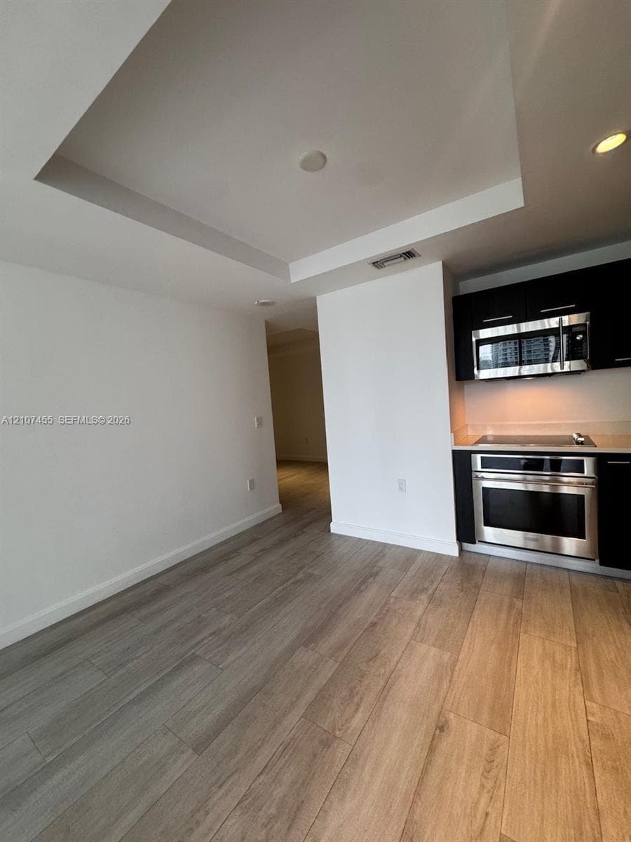 Open-concept interior featuring dark-finish custom cabinetry, integrated refrigeration, wood-finish flooring, recessed ceiling detailing, and floor-to-ceiling glass sliding doors