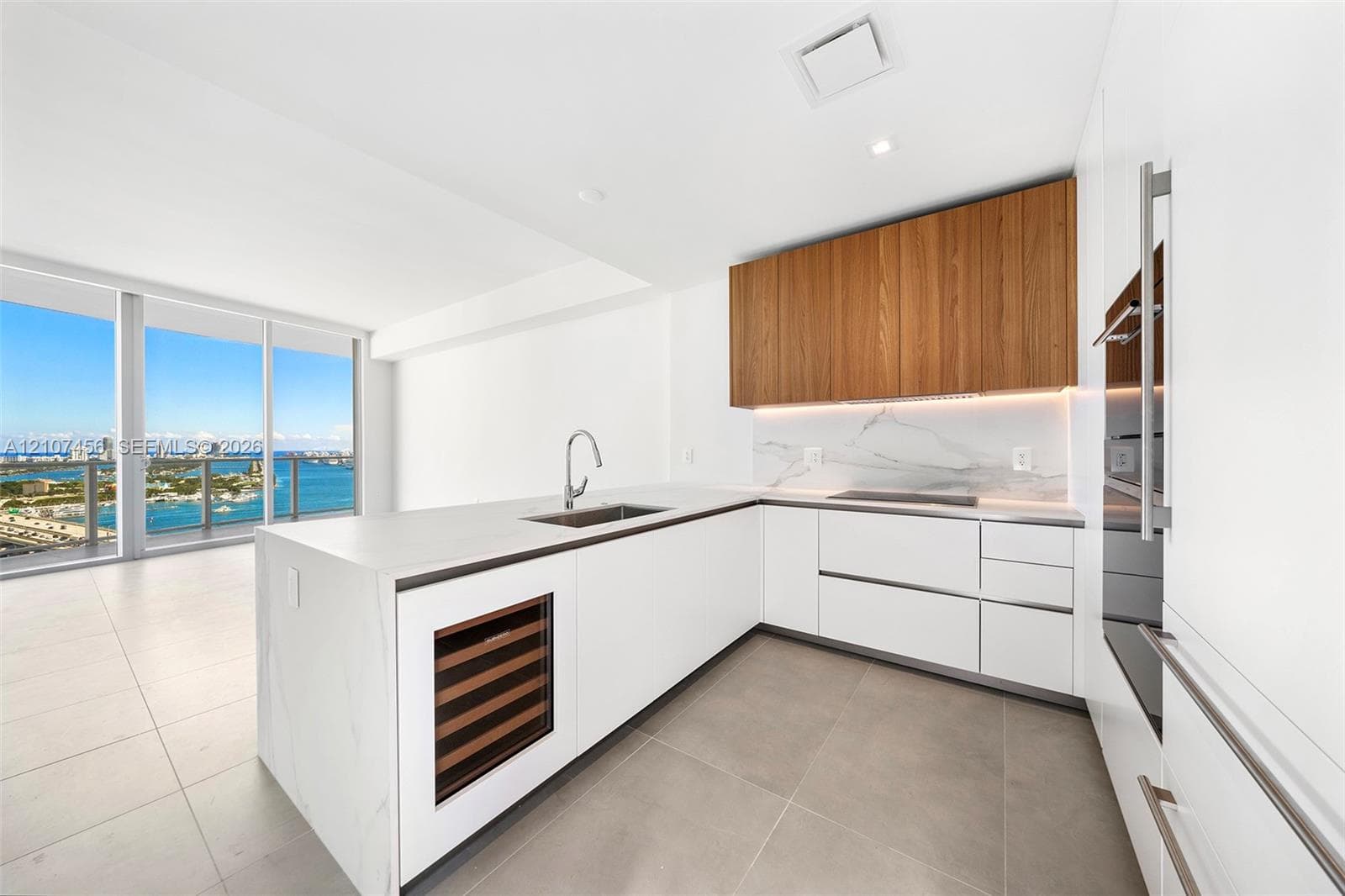 Kitchen featuring flat-panel white cabinetry, wood-finish upper cabinets, stone-surfaced island with integrated wine refrigeration, under-cabinet lighting, and floor-to-ceiling windows