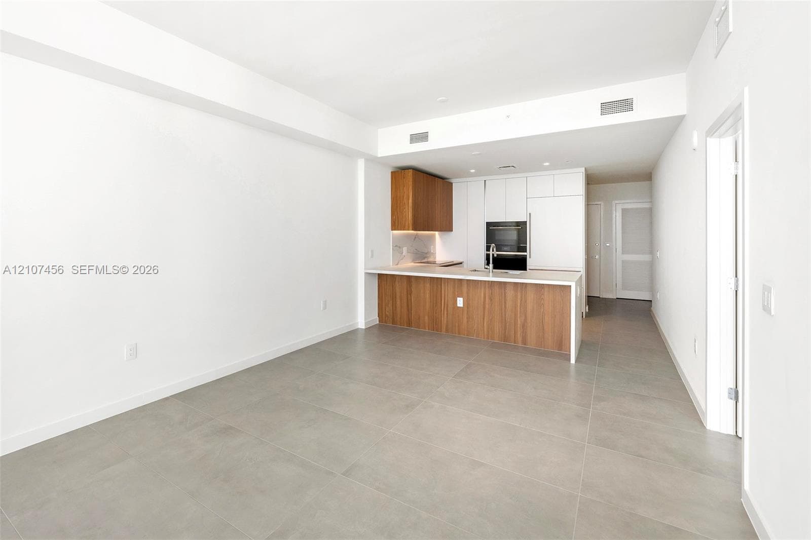 Kitchen featuring wood-finish lower cabinetry, white upper cabinets, stone-surfaced countertops, a stainless steel undermount sink, and large-format tile flooring