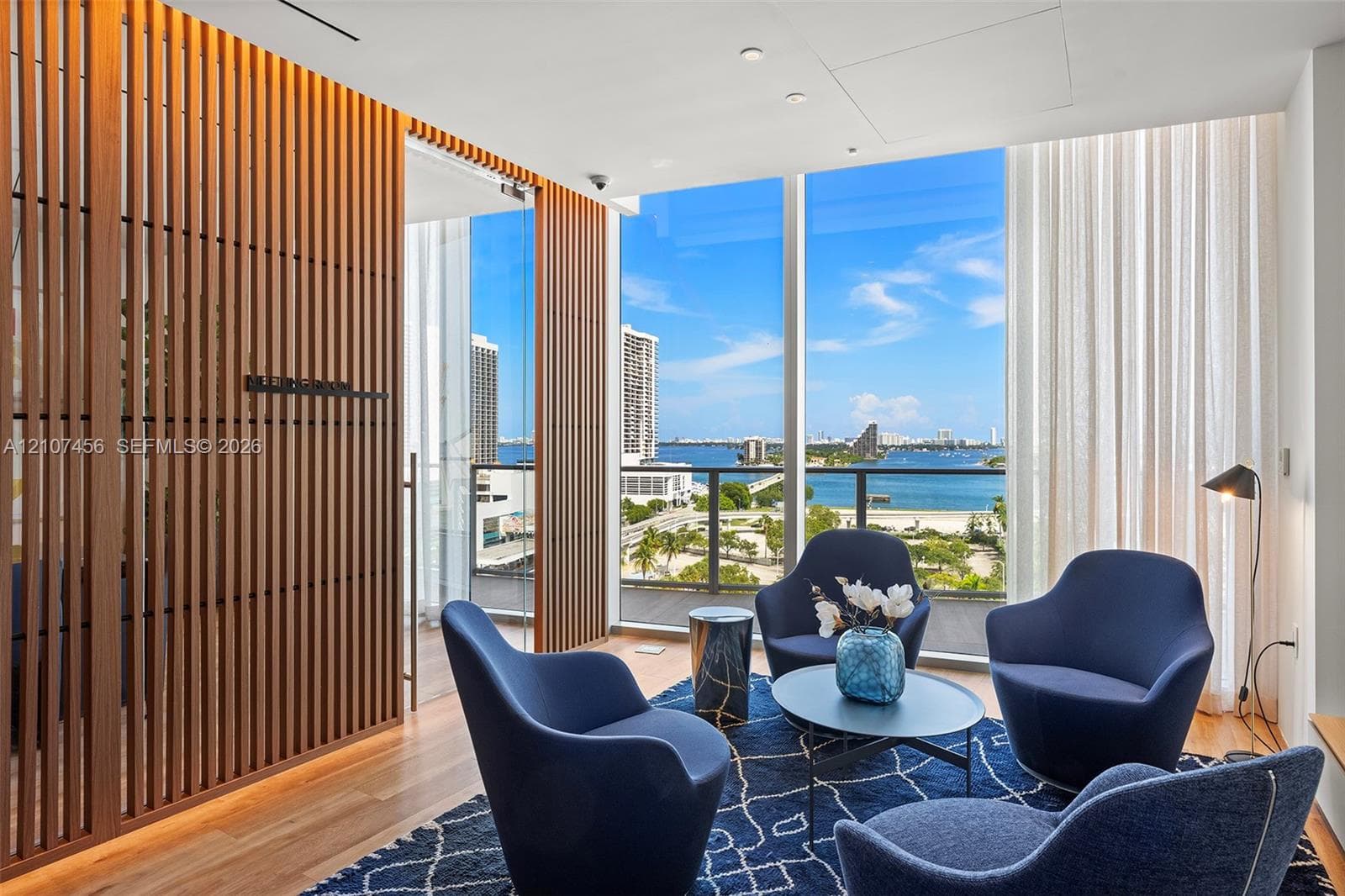 Floor-to-ceiling windows, vertical wood-slat privacy screening, recessed lighting, and a glass-railing balcony featuring a coastal waterfront backdrop