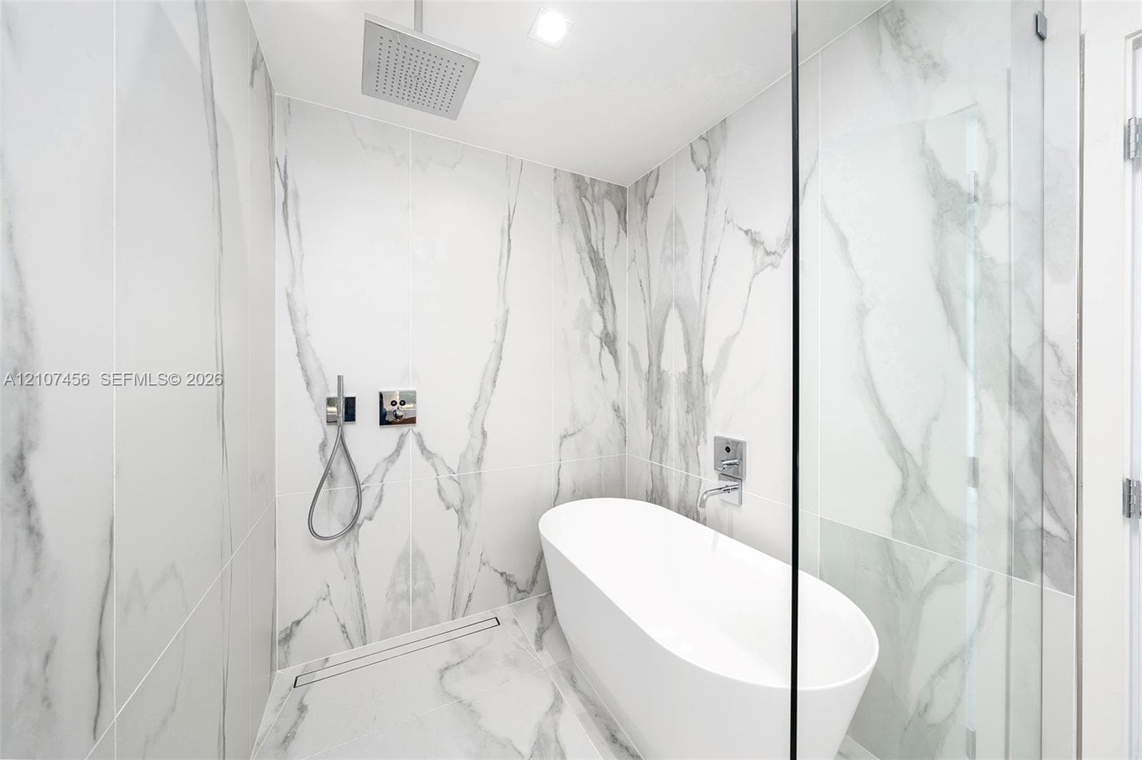 Wet room featuring floor-to-ceiling veined stone-style tile, a freestanding soaking tub, a ceiling-mounted rainfall showerhead, a wall-mounted hand-held sprayer, and a glass partition
