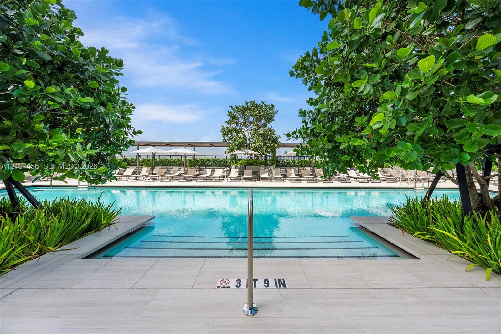 Swimming pool with stone-finish decking, stainless steel handrail, integrated steps, and surrounding mature landscaping