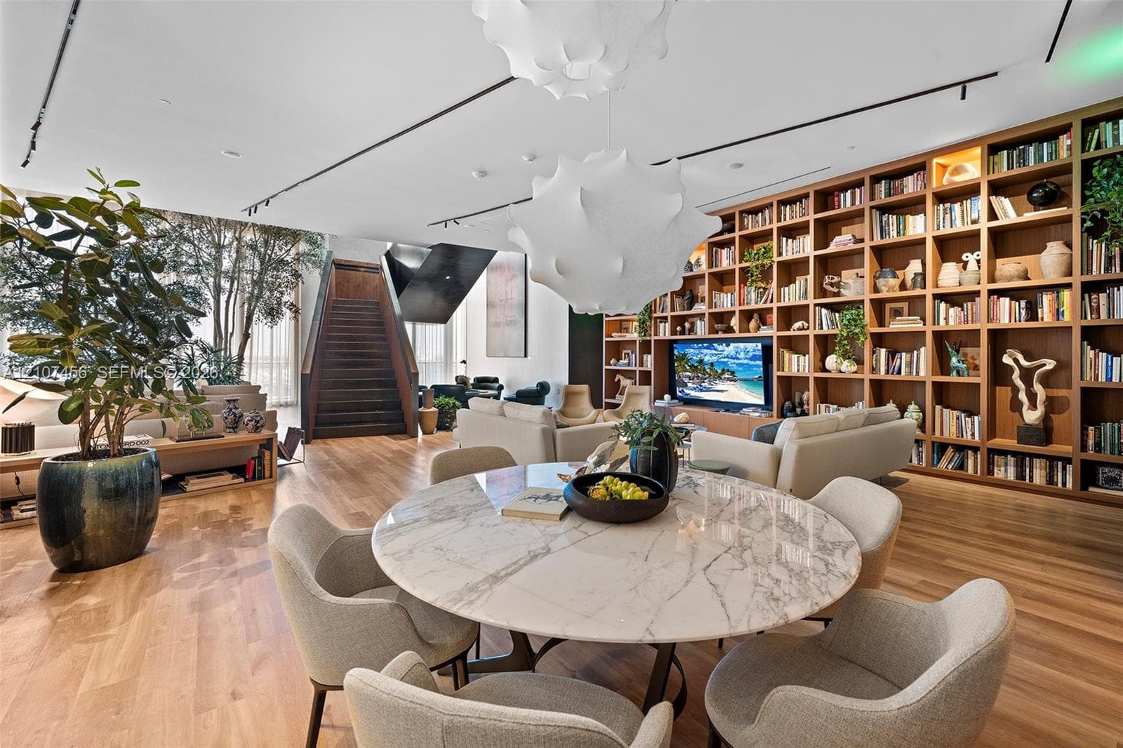 Open-concept living space featuring floor-to-ceiling built-in wooden shelving, track lighting, sculptural pendant lighting, wood-finish flooring, and a bespoke staircase
