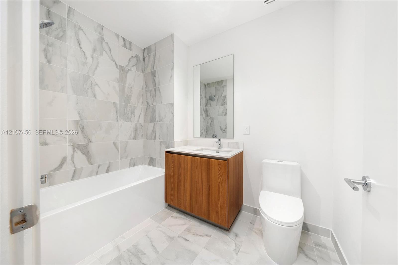 Bathroom featuring wood-finish vanity cabinetry, integrated sink with chrome faucet, wall-mounted mirror, soaking tub with tile surround, and tile flooring
