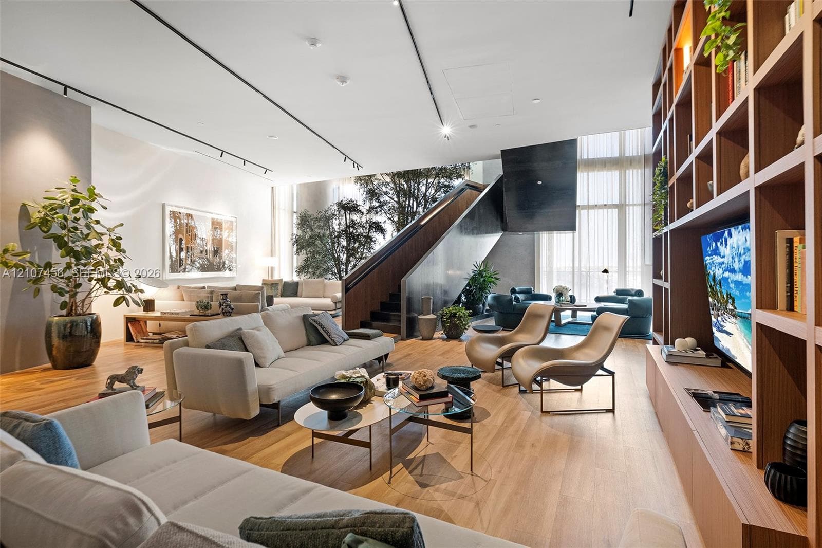 Open-concept living space featuring wood-finish flooring, floor-to-ceiling windows, custom built-in wooden shelving, a dramatic sculptural staircase, and ceiling-mounted track lighting