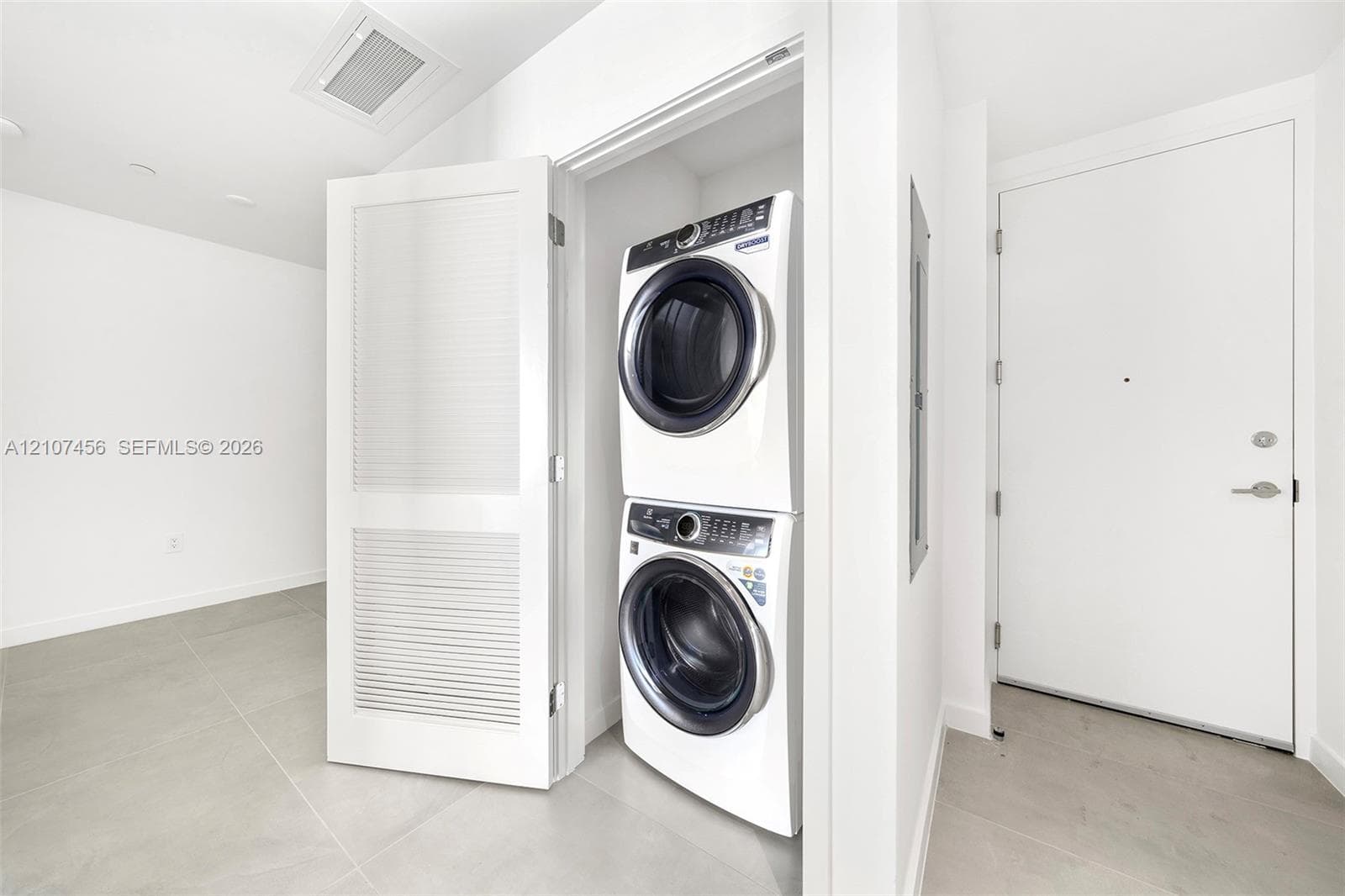 Stacked front-load laundry unit housed within a dedicated closet featuring louvered doors and large-format neutral floor tile