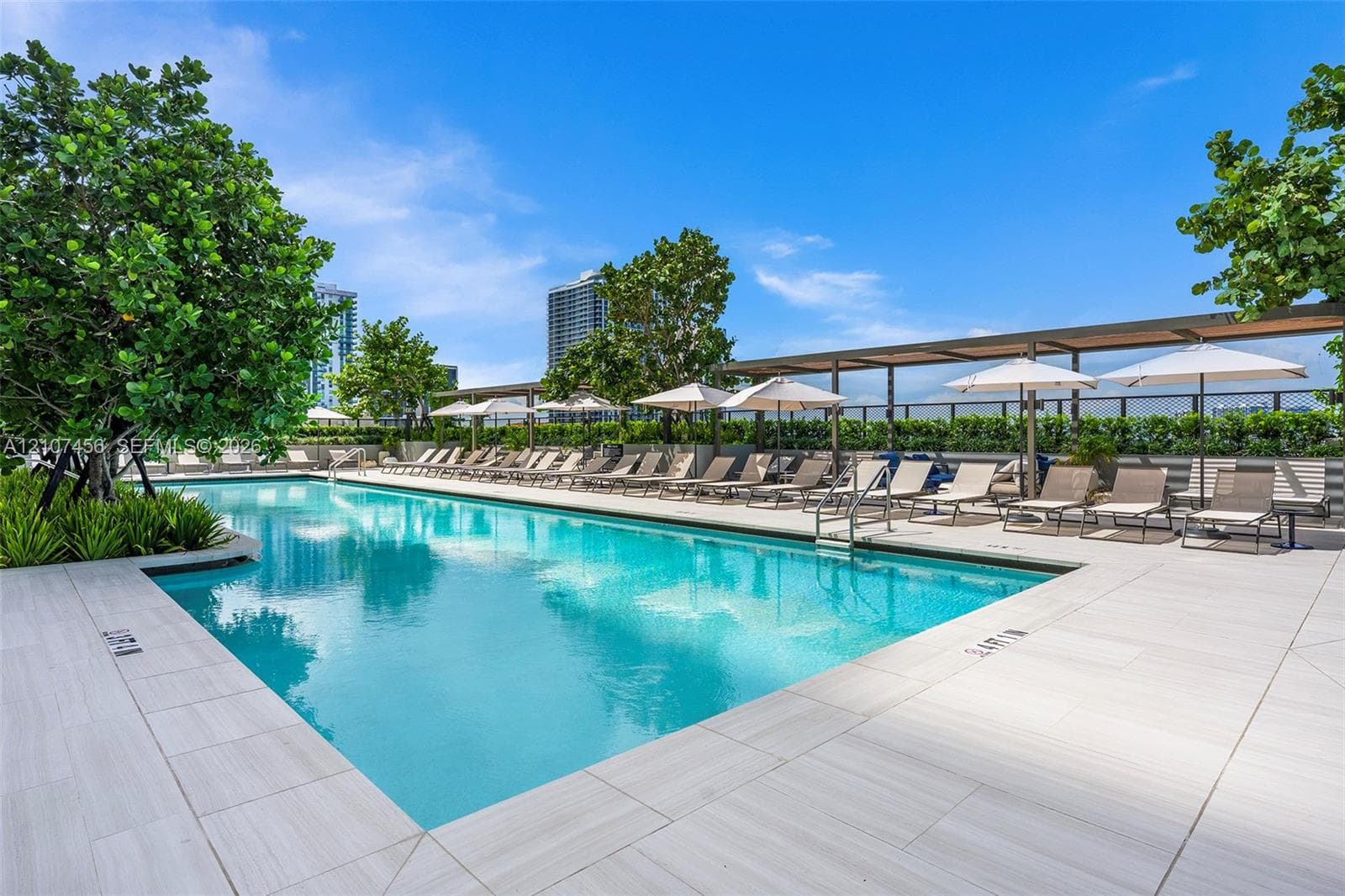 Rectangular swimming pool featuring light-toned tile decking, a metal pergola structure, and landscaped greenery
