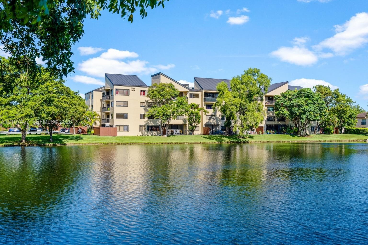 Multi-story residential building featuring a light-toned stucco exterior, pitched metal roofing, private balconies, and mature landscaping along a waterfront setting