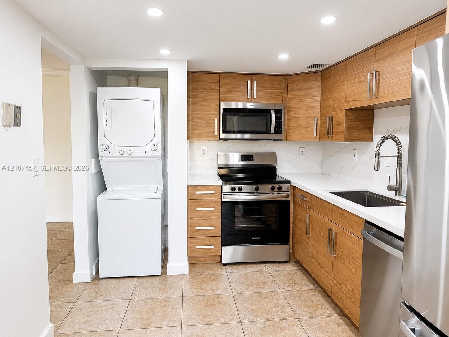 Kitchen featuring wood-finish cabinetry, stainless steel appliances, white stone-composite countertops, tile flooring, and an integrated laundry closet with a stackable washer and dryer