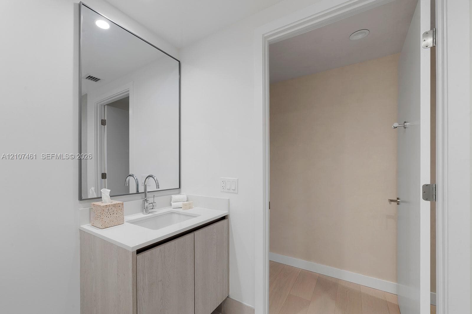 Bathroom vanity featuring light-toned cabinetry, a solid surface countertop with an integrated rectangular sink, a polished chrome bridge faucet, and a large rectangular mirror with a thin metallic frame