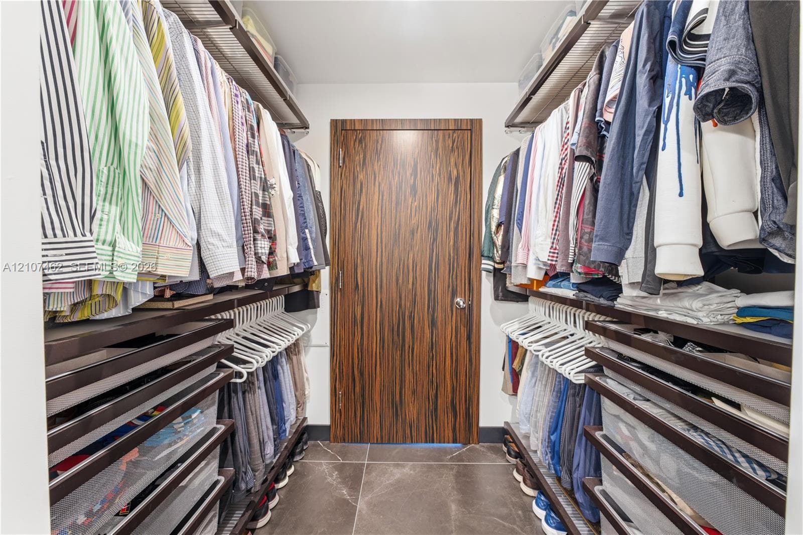 Walk-in closet featuring dual-sided integrated shelving systems, multi-tier hanging rods, pull-out storage drawers, and large-format tile flooring with a wood-finish interior door