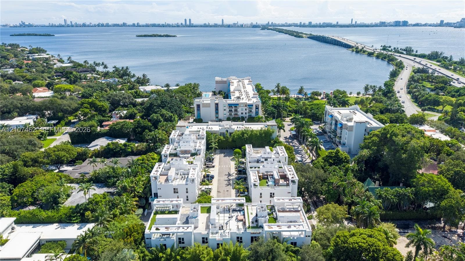 Aerial perspective of a multi-structure residential complex featuring white stucco facades, flat roofs, private rooftop terraces, and mature perimeter landscaping, situated along a coastal waterfront