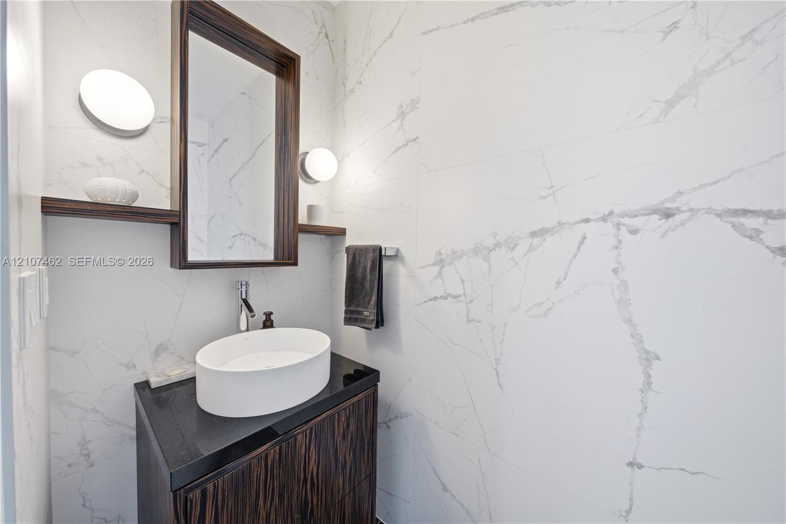 Dark wood-finish vanity with a stone countertop, white oval vessel sink, wall-mounted mirror with integrated shelving, and floor-to-ceiling stone-style wall tile