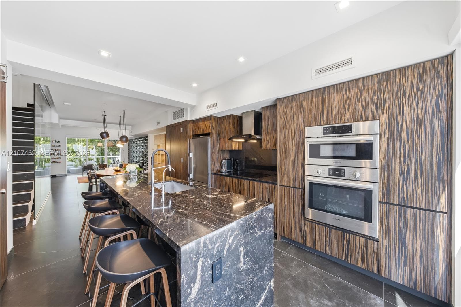 Kitchen featuring wood-finish flat-panel cabinetry, stone-surfaced waterfall island, stainless steel wall-mounted oven and microwave, and large-format tile flooring