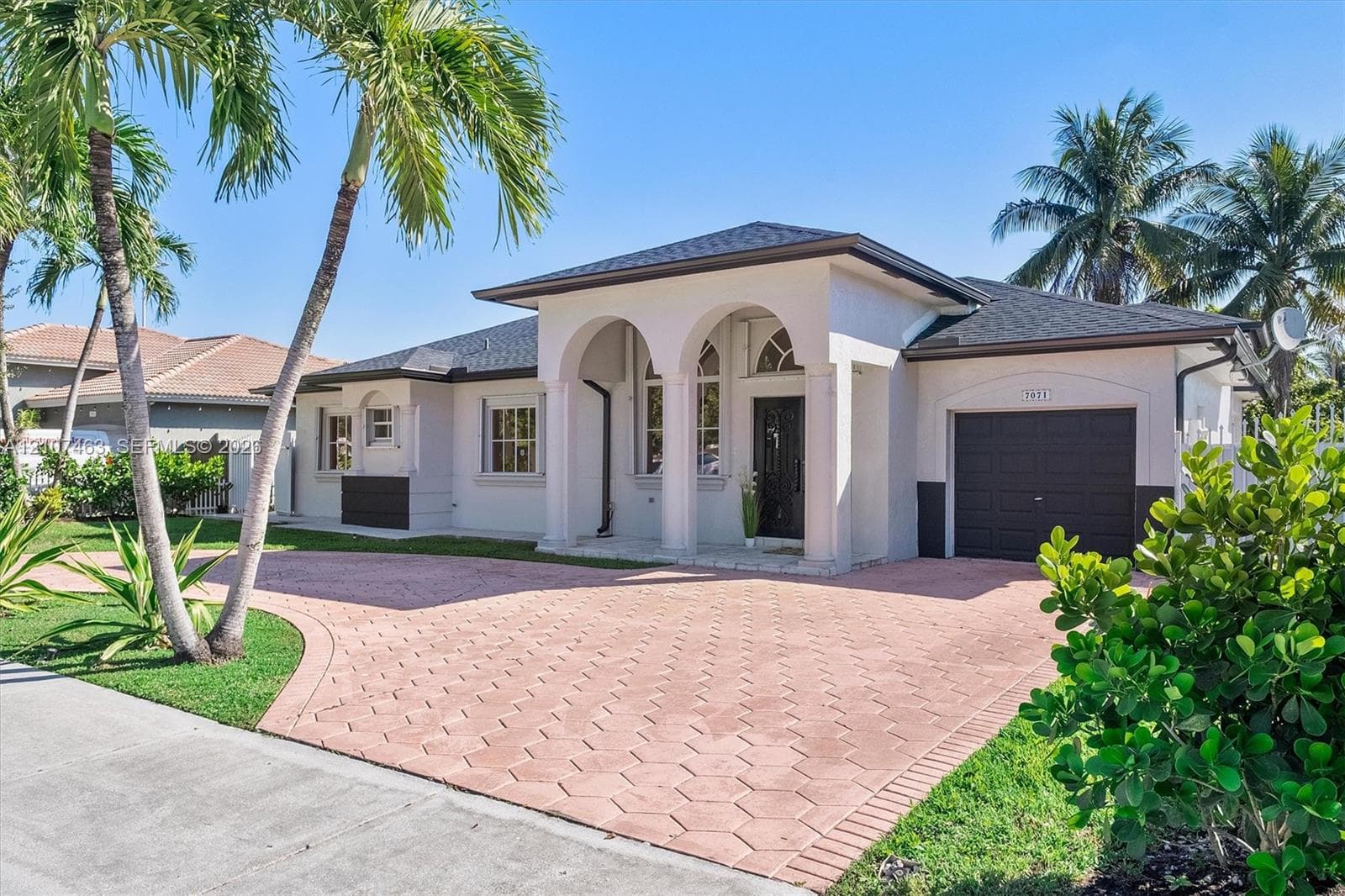 Single-story exterior featuring a covered arched entryway, hexagonal paved driveway, dark-toned garage door, and mature palm trees