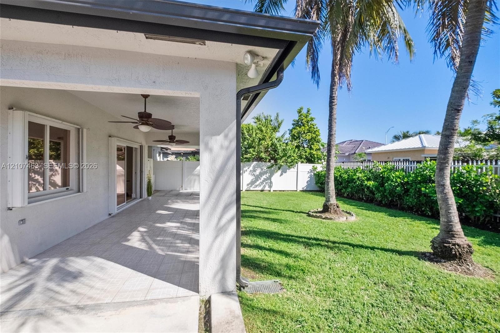 Covered concrete patio featuring ceiling-mounted fans and sliding glass doors, overlooking a lawn enclosed by white vinyl fencing and palm trees