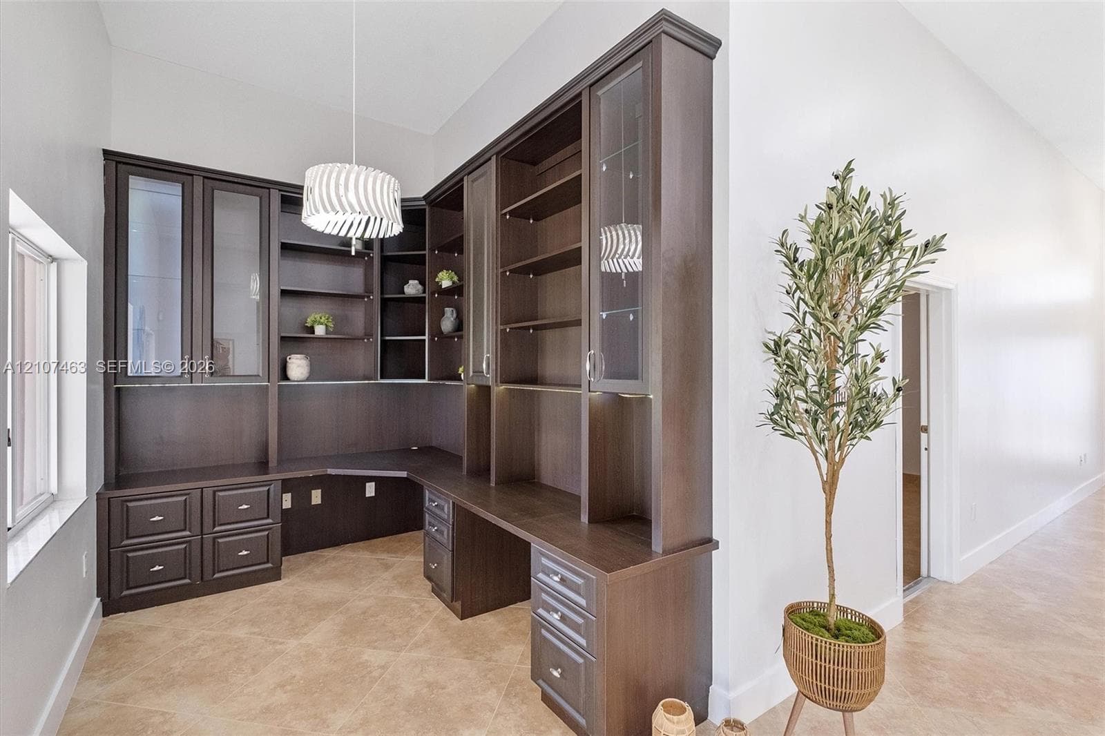 Built-in dark wood cabinetry featuring glass-front display doors, open shelving, integrated desk surface, and drawer storage, complemented by high ceilings and neutral tile flooring