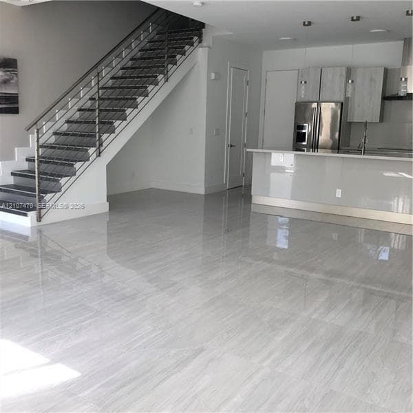 Open-concept interior featuring large-format tile flooring, a modern staircase with a metal cable railing, flat-panel cabinetry, a kitchen island with a stone-finish countertop, and stainless steel appliances
