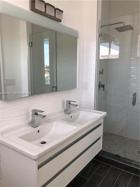 Double vanity with rectangular undermount sinks, polished chrome faucets, textured wall tile backsplash, frameless glass shower enclosure, and wood-finish flooring