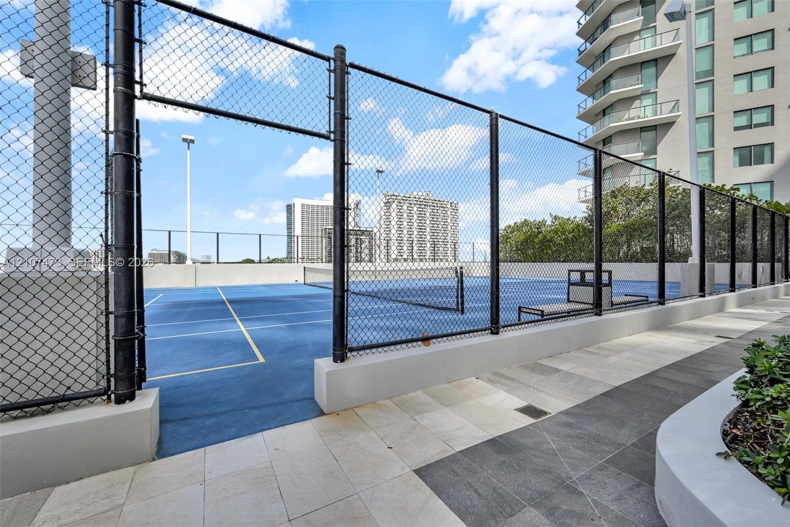 Tennis court featuring a blue hard surface, perimeter chain-link fencing, built-in court seating, and stone-tiled walking paths
