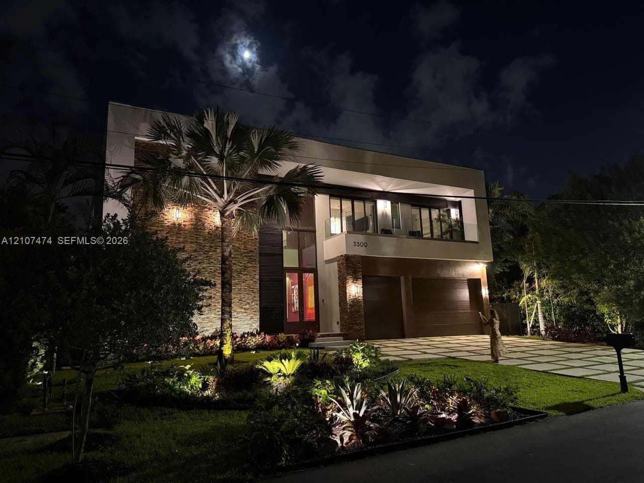 Modern two-story architectural exterior featuring stone facade detailing, integrated landscape lighting, glass-railed upper balcony, and a multi-car garage with wood-finish doors