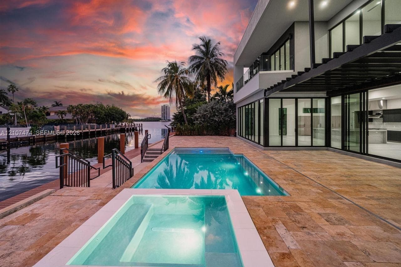 Contemporary waterfront residence featuring an in-ground pool and integrated spa, expansive stone-tiled patio, private dock, and floor-to-ceiling glass wall systems