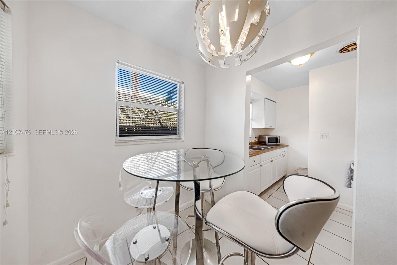 Dining area featuring a modern sculptural pendant light fixture, ceramic tile flooring, and a window with blinds, opening into a kitchen equipped with white cabinetry and stone countertops