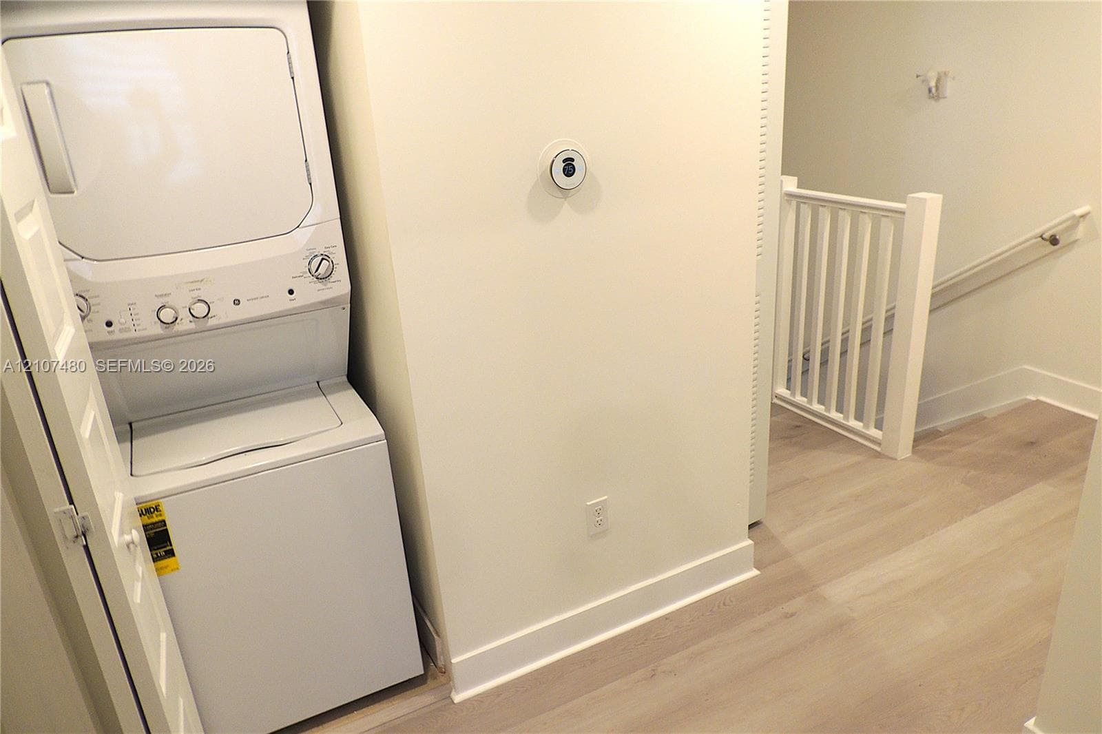 Laundry area featuring a stacked washer and dryer unit, wood-finish flooring, smart thermostat, and a white balustrade staircase