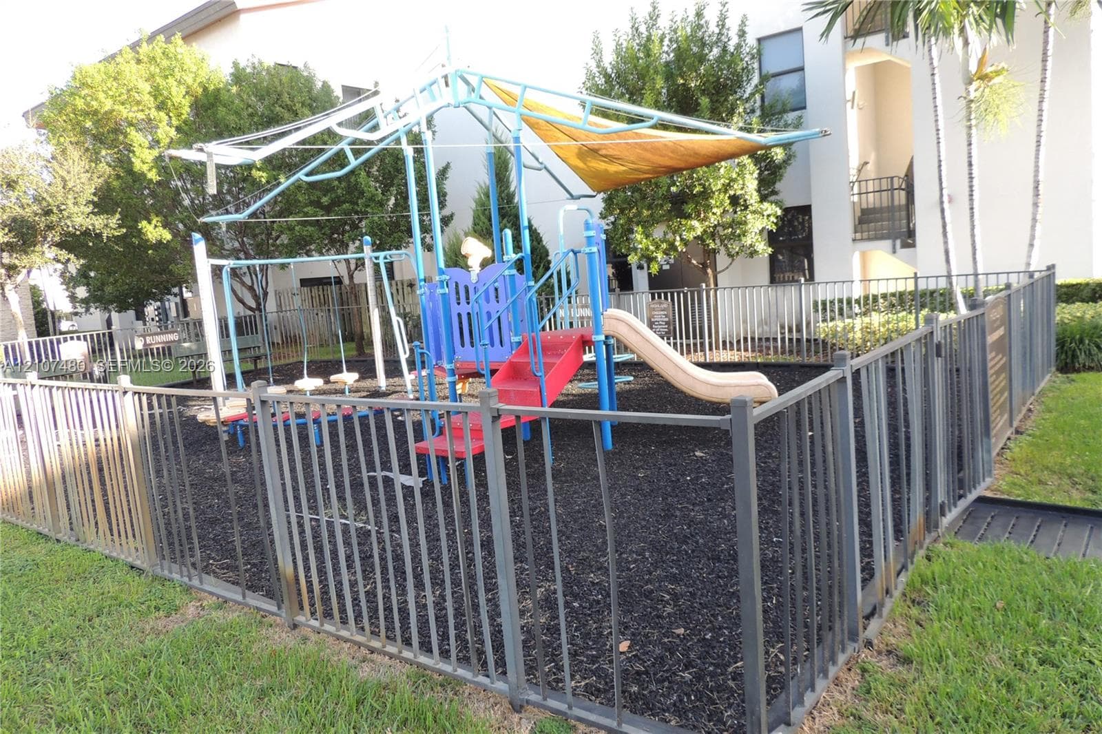 Recreational area featuring a playground structure with a shade canopy, slide, and climbing elements, enclosed by metal fencing with ground-level mulch surfacing