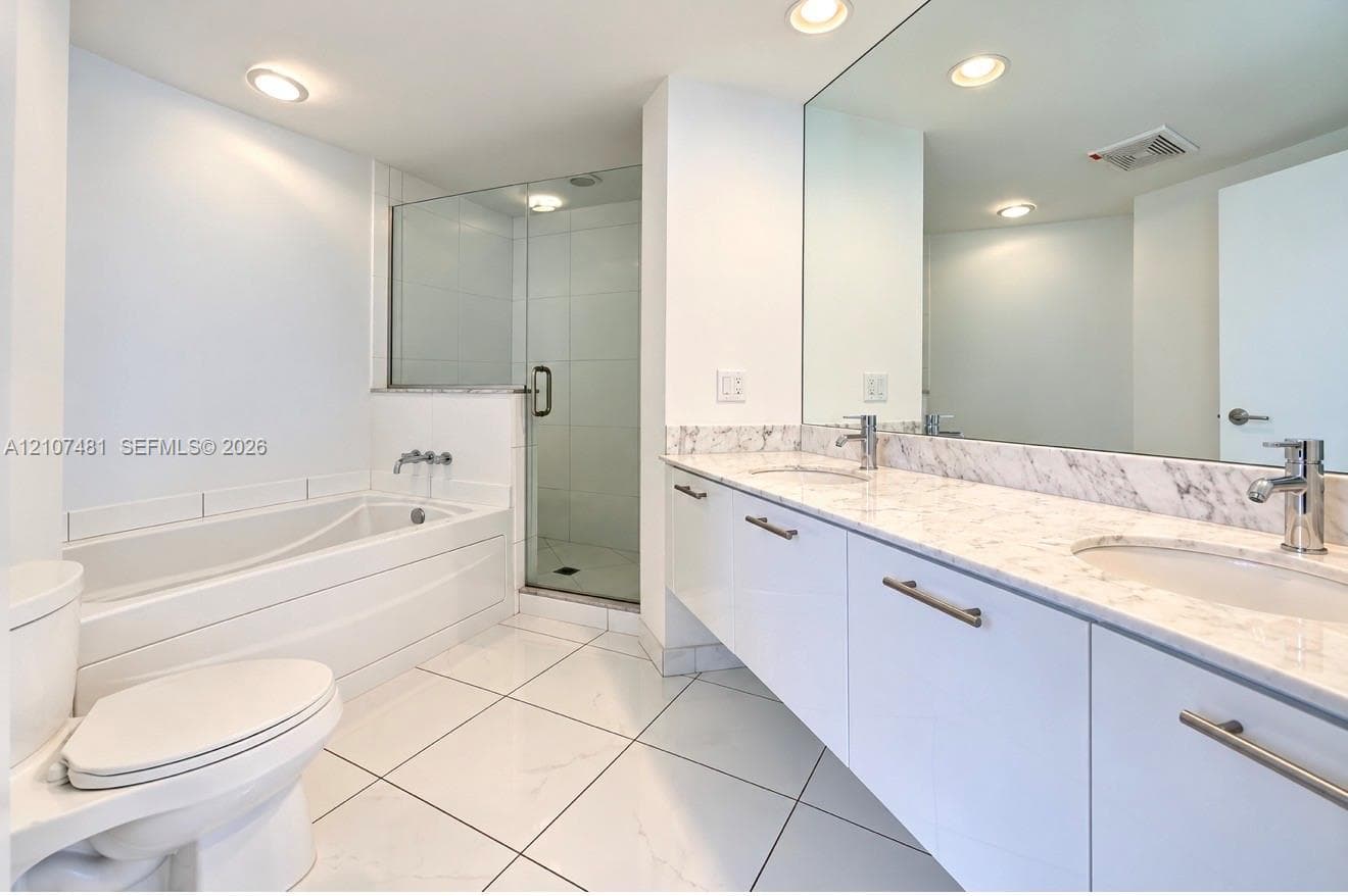 Floating vanity with dual rectangular undermount sinks, stone-finish countertops, glass-enclosed shower, deep soaking tub, and polished tile flooring