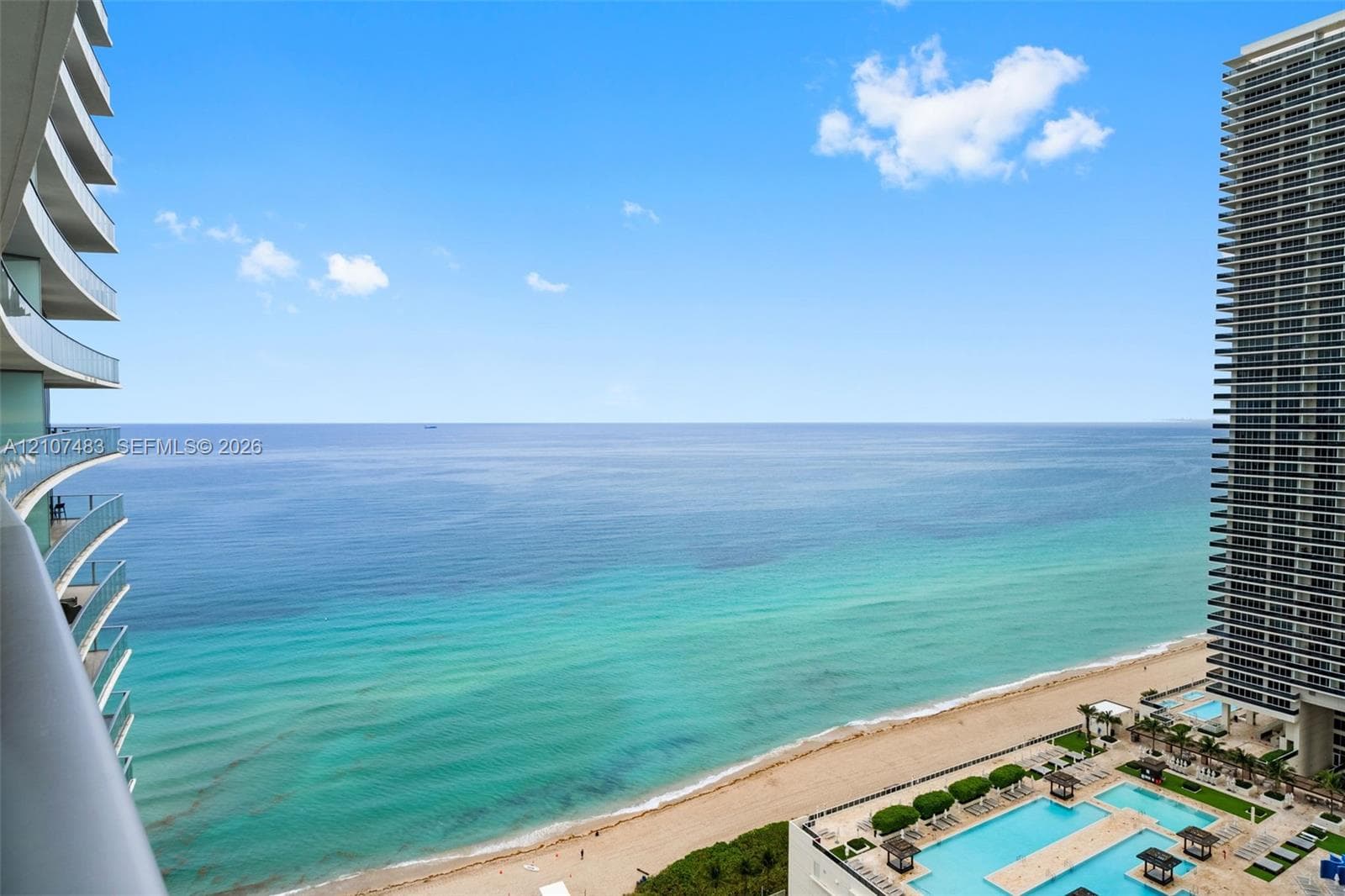 Private balcony featuring glass railings, overlooking the coastline, sandy beach, and multi-level outdoor swimming pools