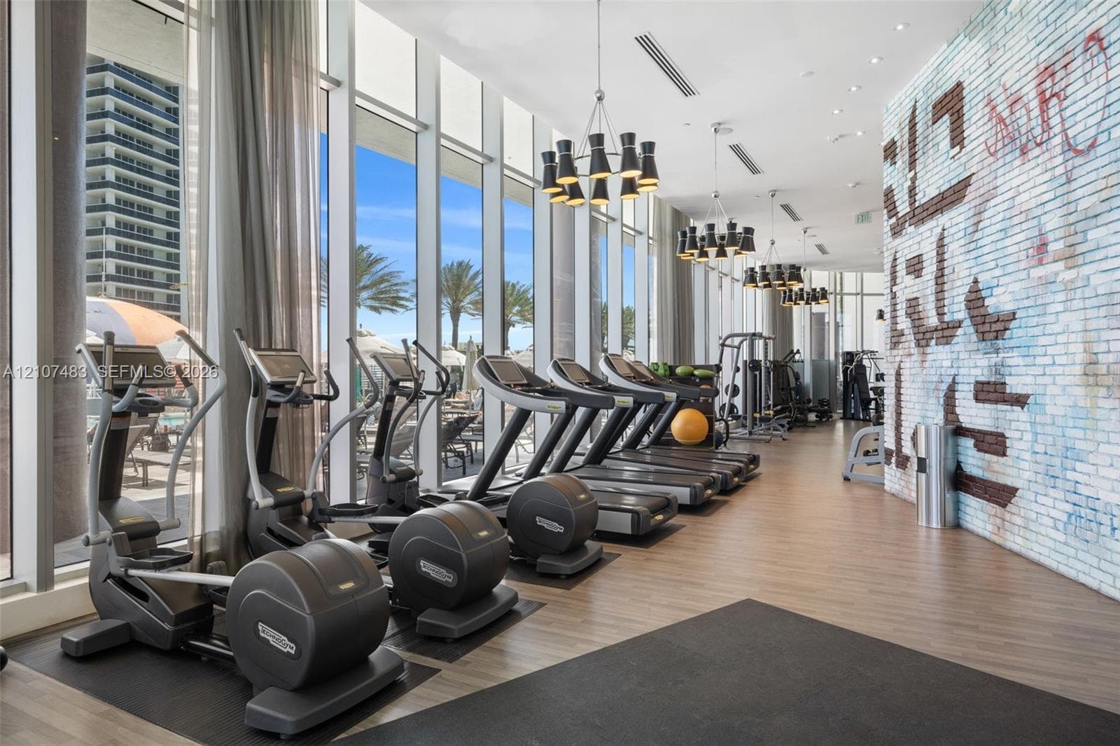 Fitness center featuring floor-to-ceiling windows, wood-finish flooring, decorative brick accent wall, and multi-tier modern chandelier lighting