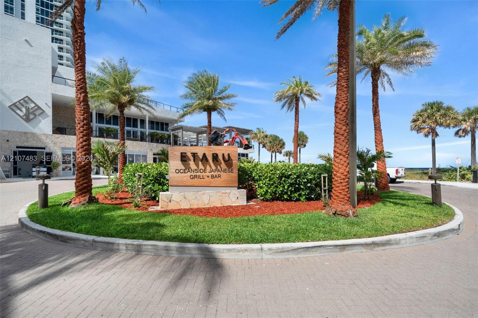 Circular paved entryway featuring mature palm trees, manicured landscaping, and natural stone signage