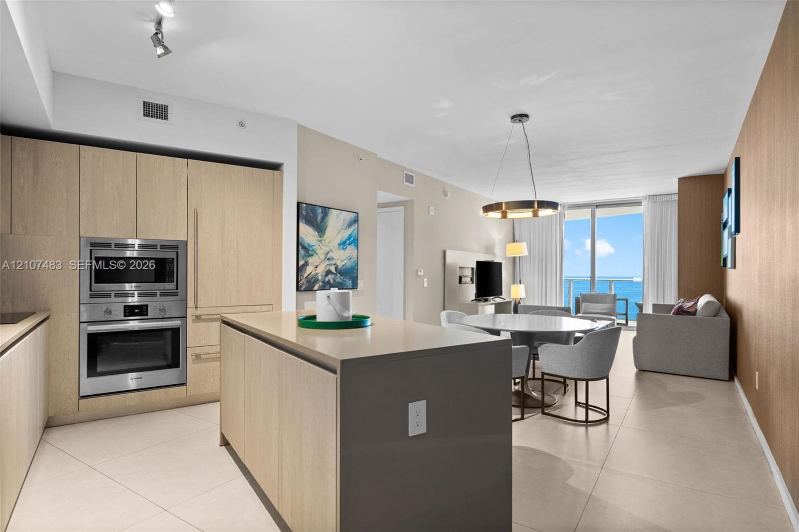 Open-concept kitchen featuring light wood-finish cabinetry, a central island with a solid surface countertop, built-in stainless steel double wall ovens, and large-format tile flooring