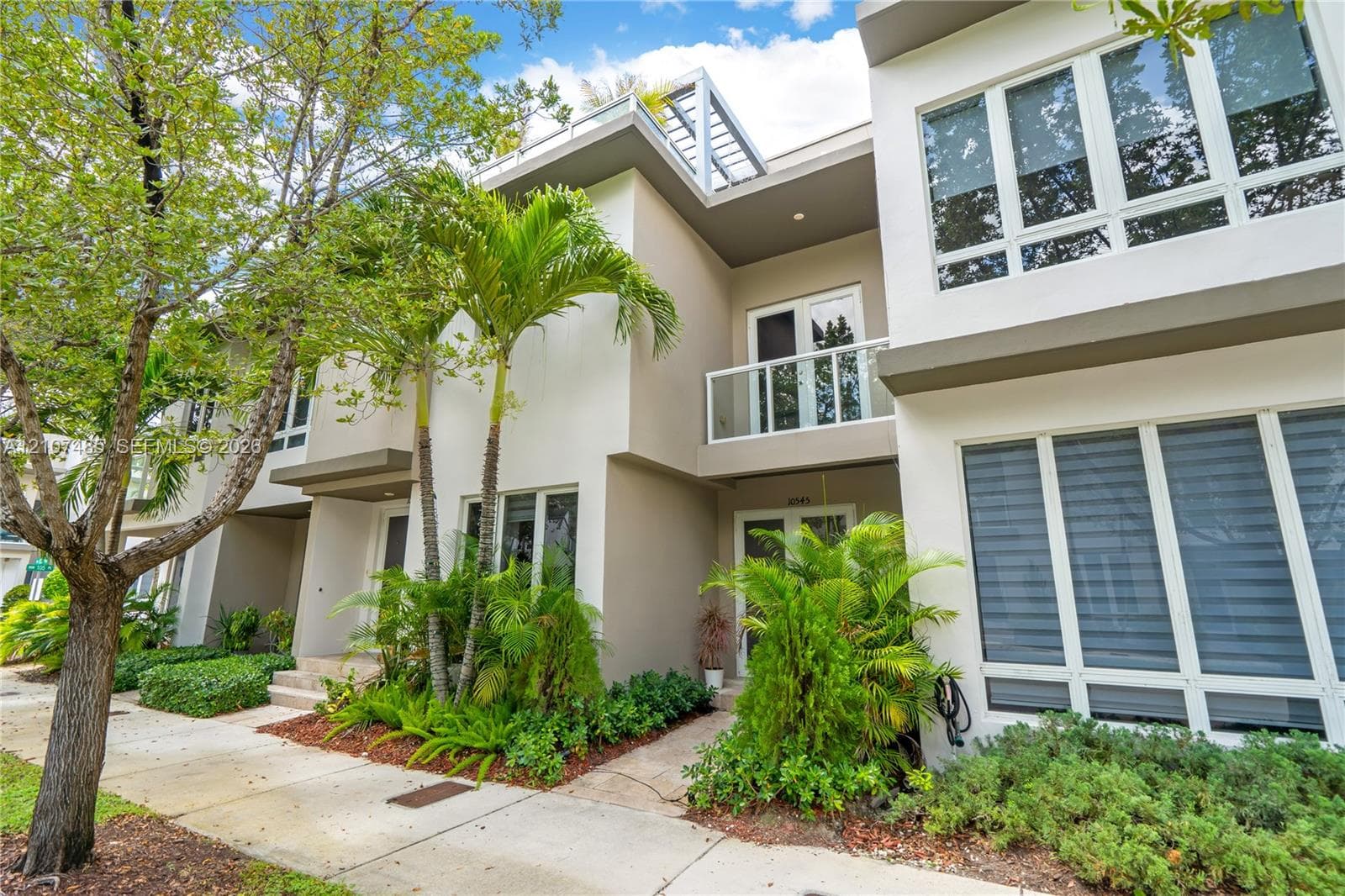 Modern multi-story exterior featuring a smooth stucco facade, expansive floor-to-ceiling windows, a private second-story balcony with glass railing, and a metal roof pergola