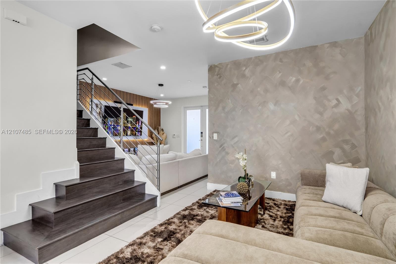 Staircase with dark wood-finish treads and metal cable railings, custom textural accent wall, circular LED chandelier, and polished tile flooring