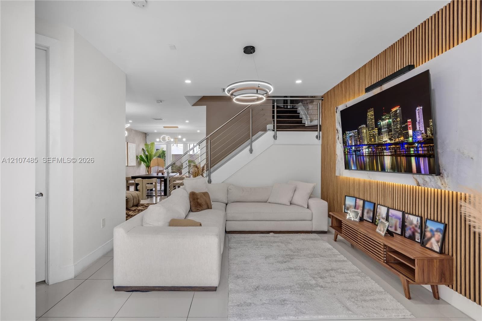 Accent wall featuring vertical wood slat paneling with integrated recessed lighting, stone-finish hearth, porcelain tile flooring, modern multi-tiered ring chandelier, and stainless steel stair railing