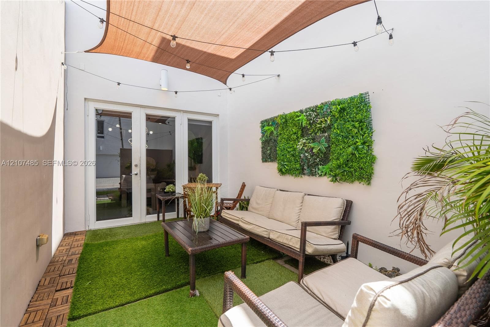 Enclosed patio area featuring white stucco walls, synthetic turf surfacing, a retractable fabric shade canopy, integrated string lighting, and a wall-mounted artificial vertical garden