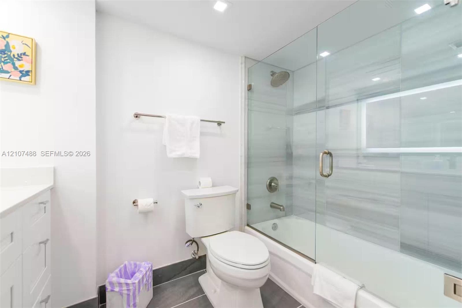 Bathroom featuring a tub-shower combination enclosed by a frameless glass door, horizontal tile wall surround, recessed lighting, white vanity cabinet with a stone countertop, and gray large-format tile flooring