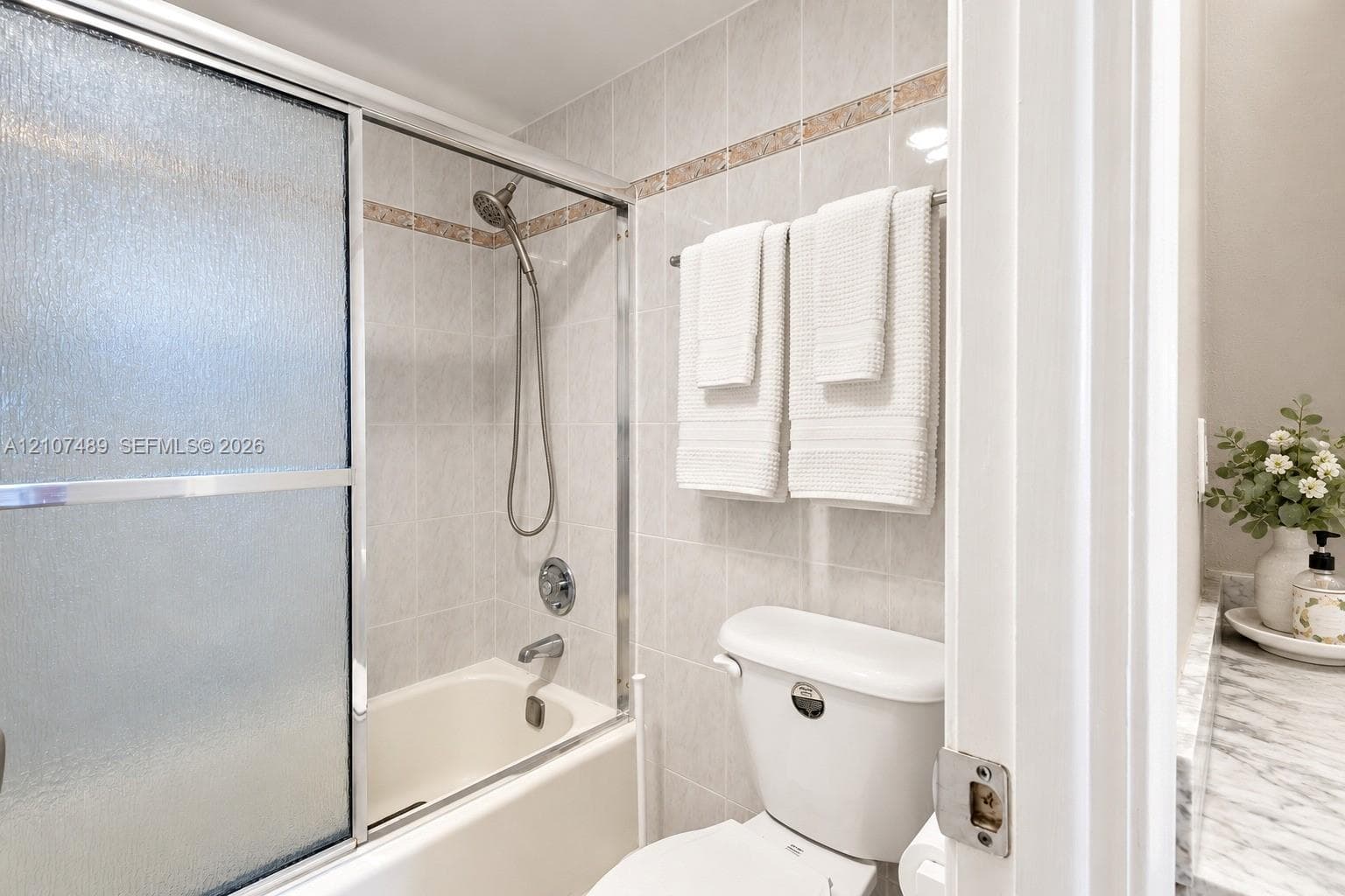 Bathtub with glass sliding doors, integrated handheld shower fixture, light-toned wall tile with decorative accent border, and stone-finish vanity countertop