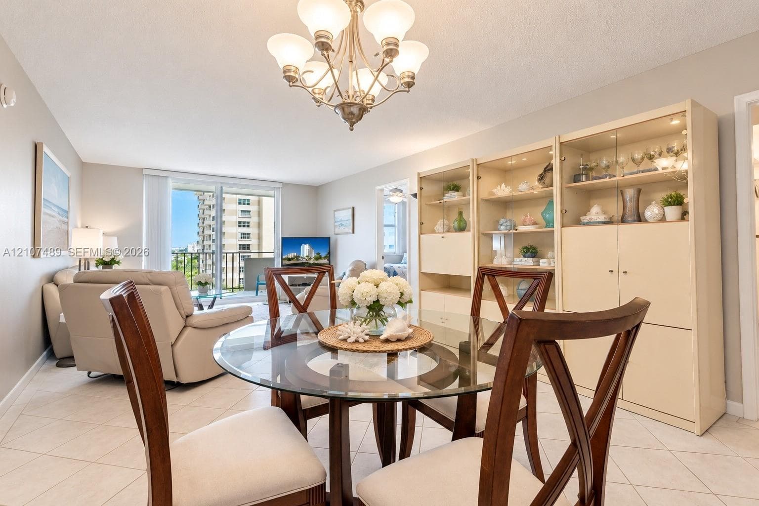Living area featuring light-toned tile flooring, a multi-bulb chandelier, built-in floor-to-ceiling cabinetry with glass shelving, and sliding glass doors providing outdoor access
