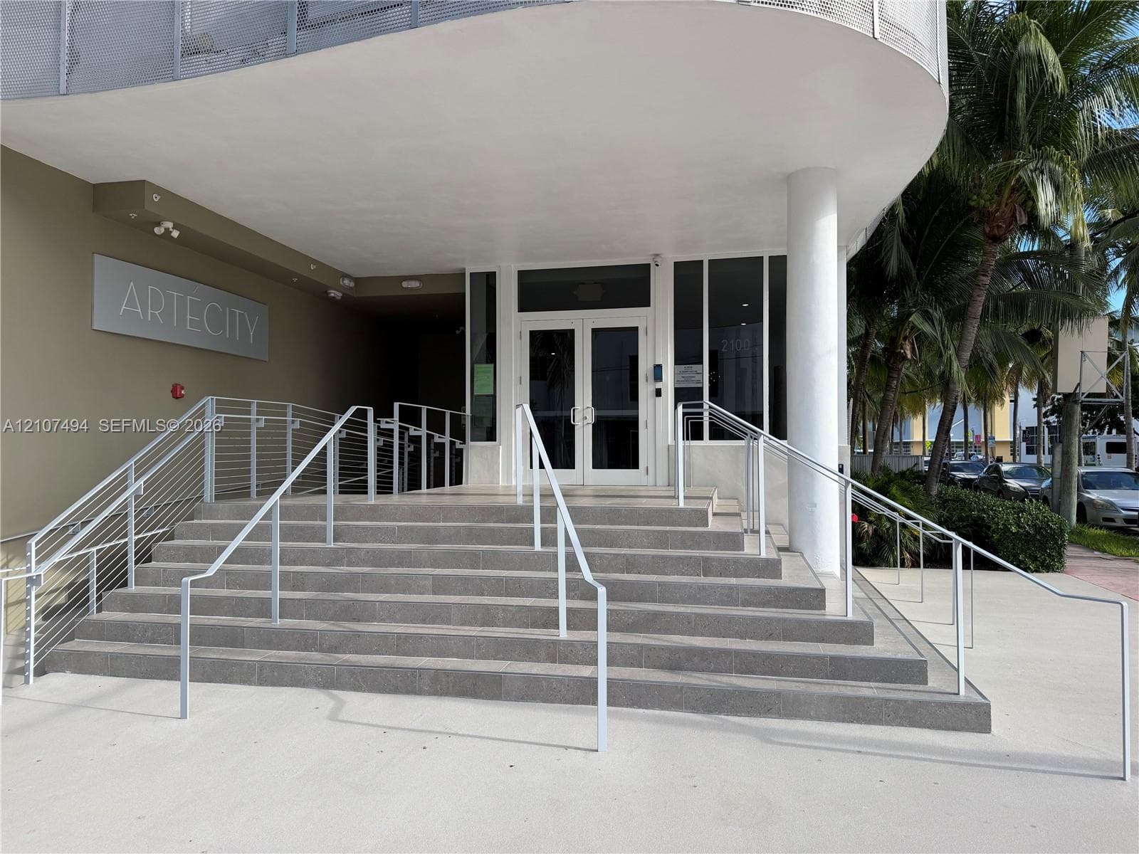 Building entrance featuring a concrete staircase with metal railings, a covered porch, glass-paned double entry doors, and a concrete-paved approach