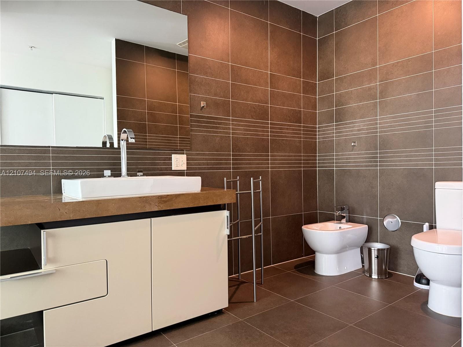 Wall-to-wall brown ceramic tile, custom white vanity with a rectangular vessel sink, polished chrome fixtures, integrated bidet, and large-format wall mirror