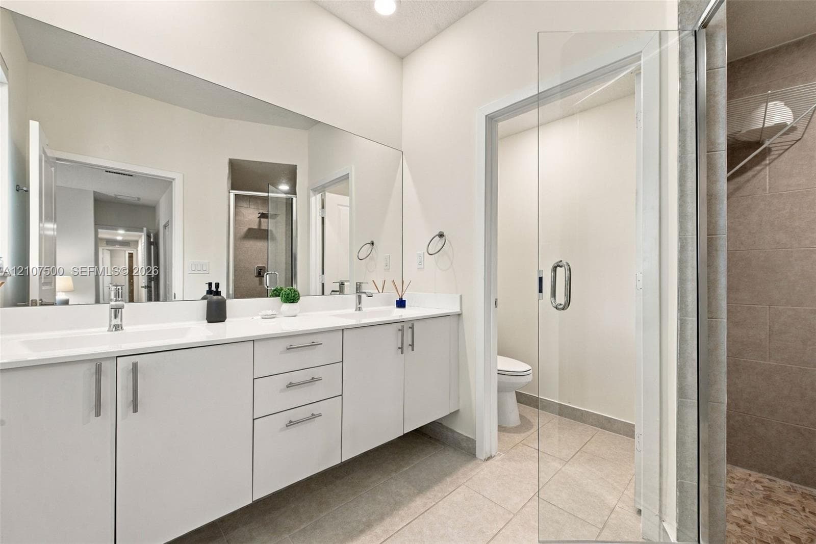 Bathroom featuring a dual-sink floating vanity with white countertops, expansive wall-to-wall mirror, glass-enclosed shower, and neutral tile flooring
