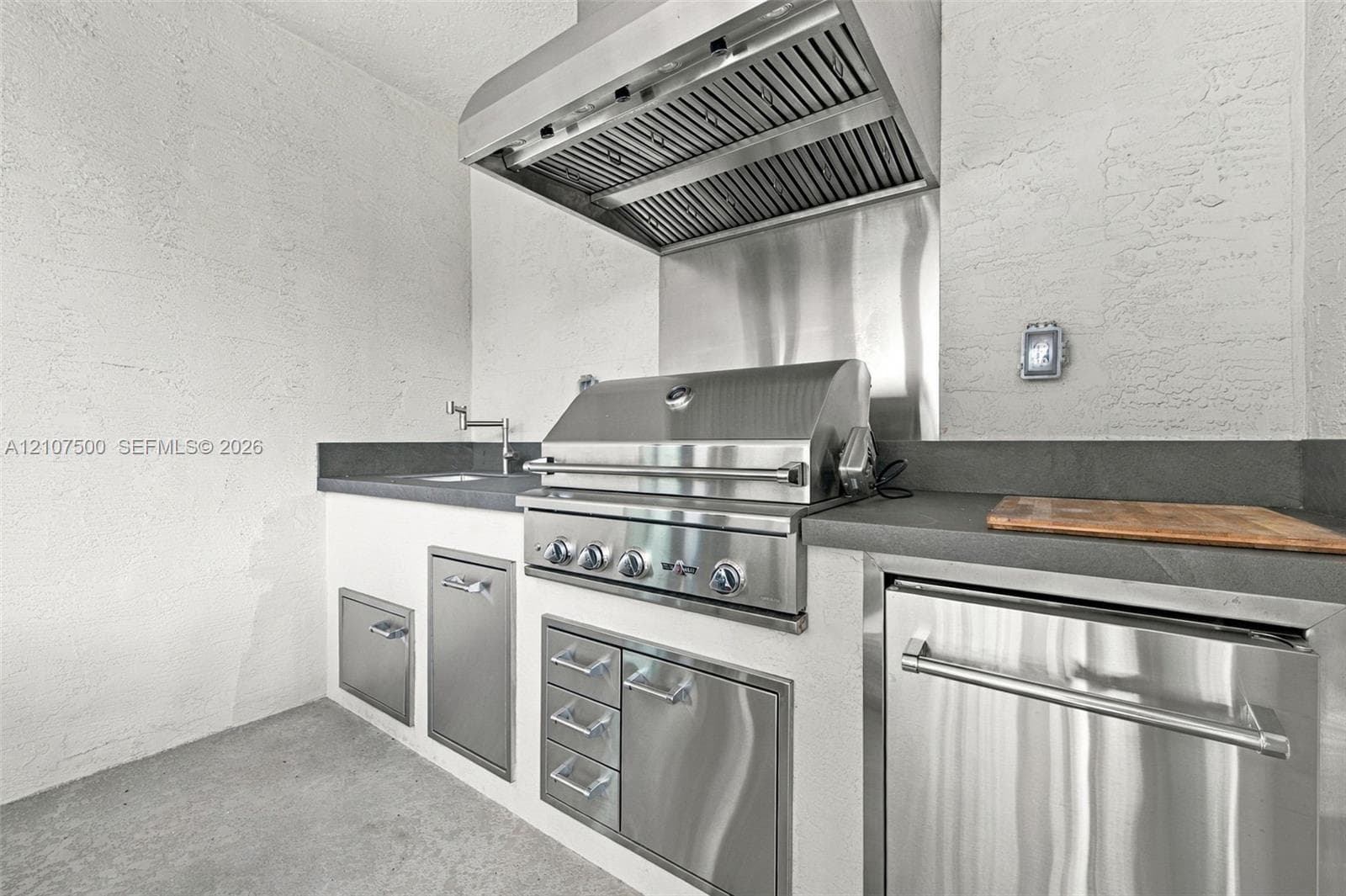 Outdoor kitchen featuring stainless steel built-in grill, professional-grade range hood, stone-finish countertops, stainless steel under-counter cabinetry, and integrated prep sink with faucet